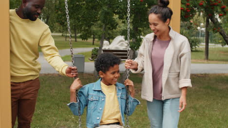 Little-African-American-Boy-Swinging-on-Swing-in-Park-with-Mother-and-Dad